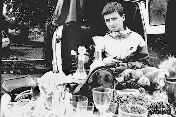 Uninspired boy waits for a customer at his car boot sale, Vallance Rd, London 1984